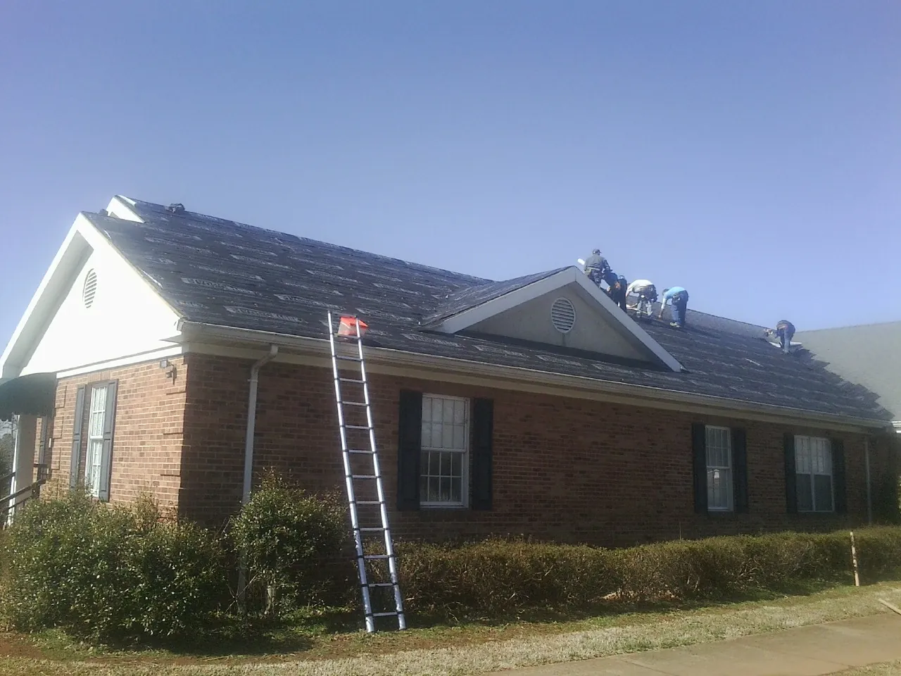Crew tearing shingles off a brick ranch roof with a ladder set — Emergency Roof Repair work by our crew in East Ellijay