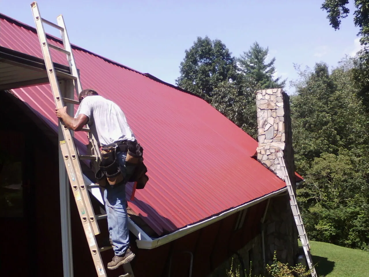 Roofer on a ladder beside a red metal roof and stone chimney — Soffit & Fascia Repair by Roofing Brothers in East Ellijay